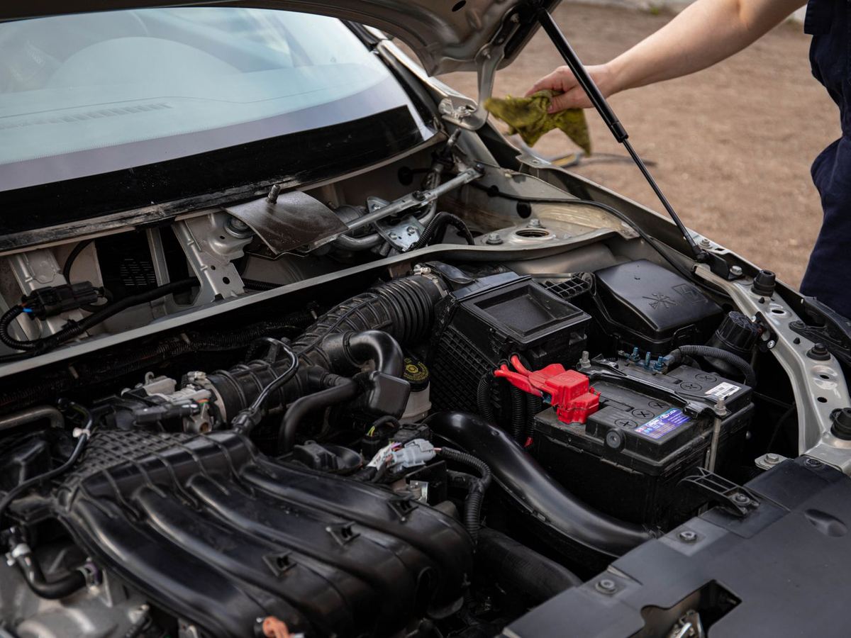 Stock photo: an open hood showing a car battery in the engine bay, with a mechanic at the side.